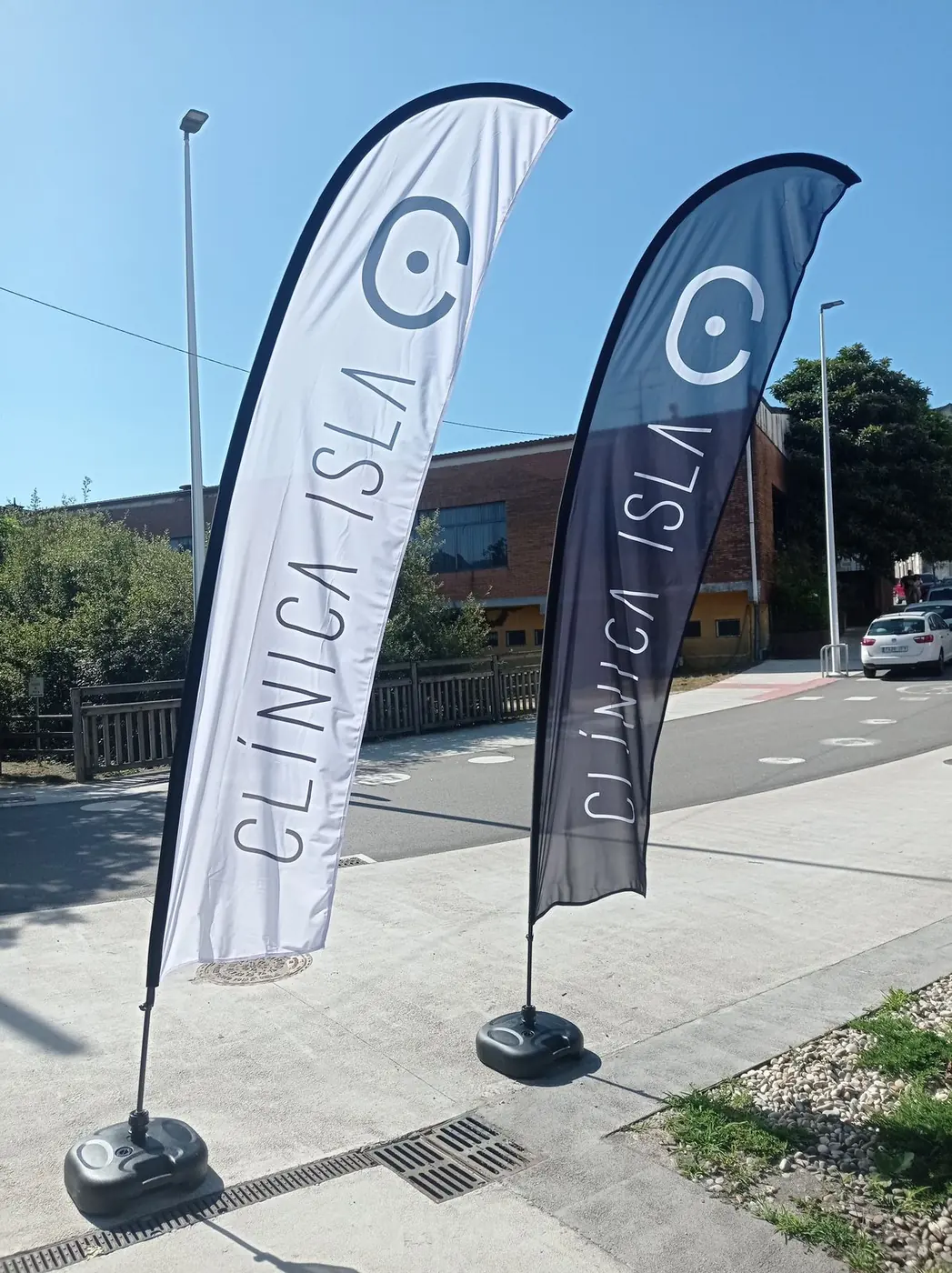 Dos banderas publicitarias de pluma de Clínica Isla, una blanca y otra gris, con su base, en la acera