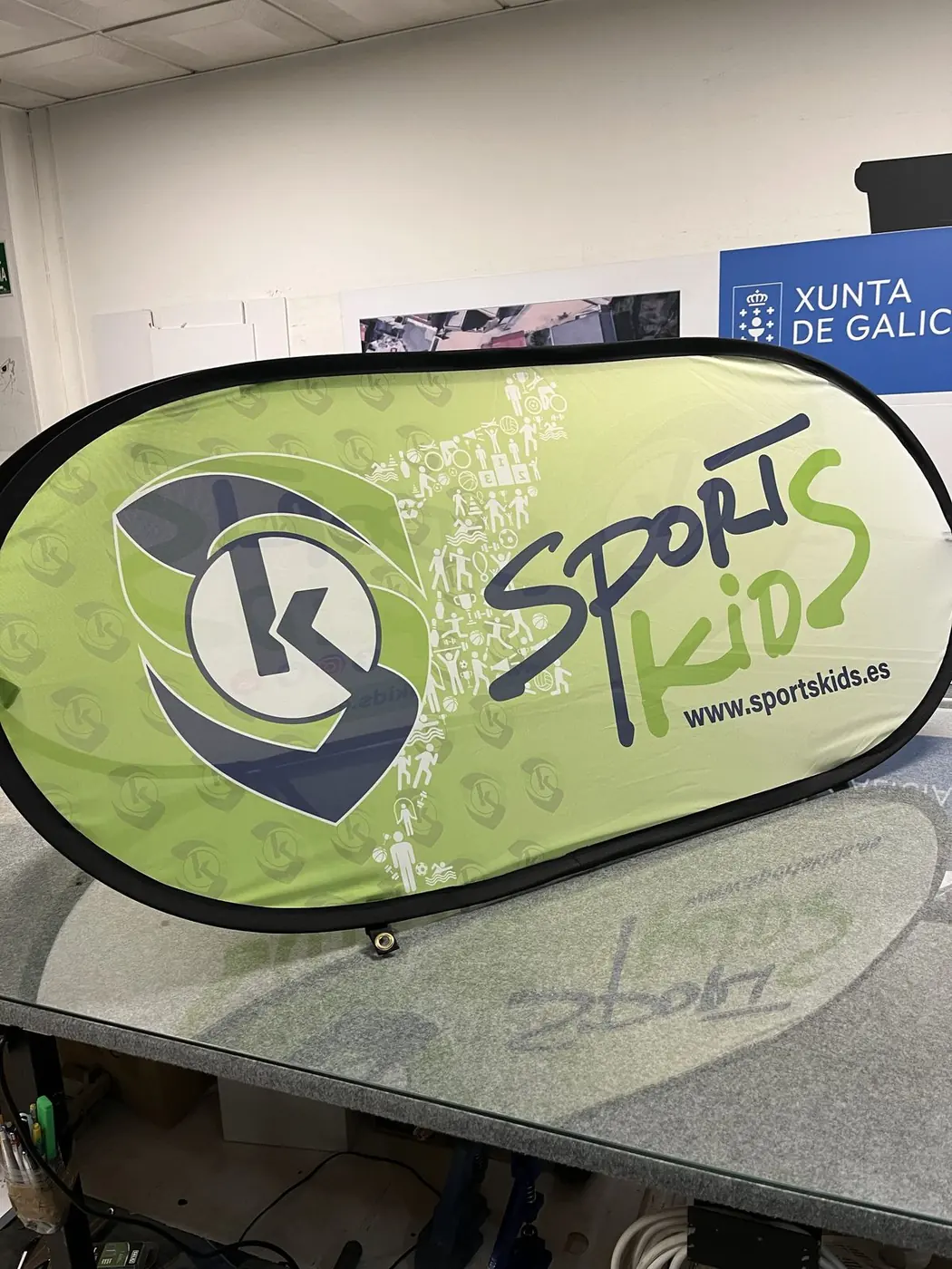 Pancarta elástica verde de Sport Kids con el logotipo y la web, sobre una mesa del taller