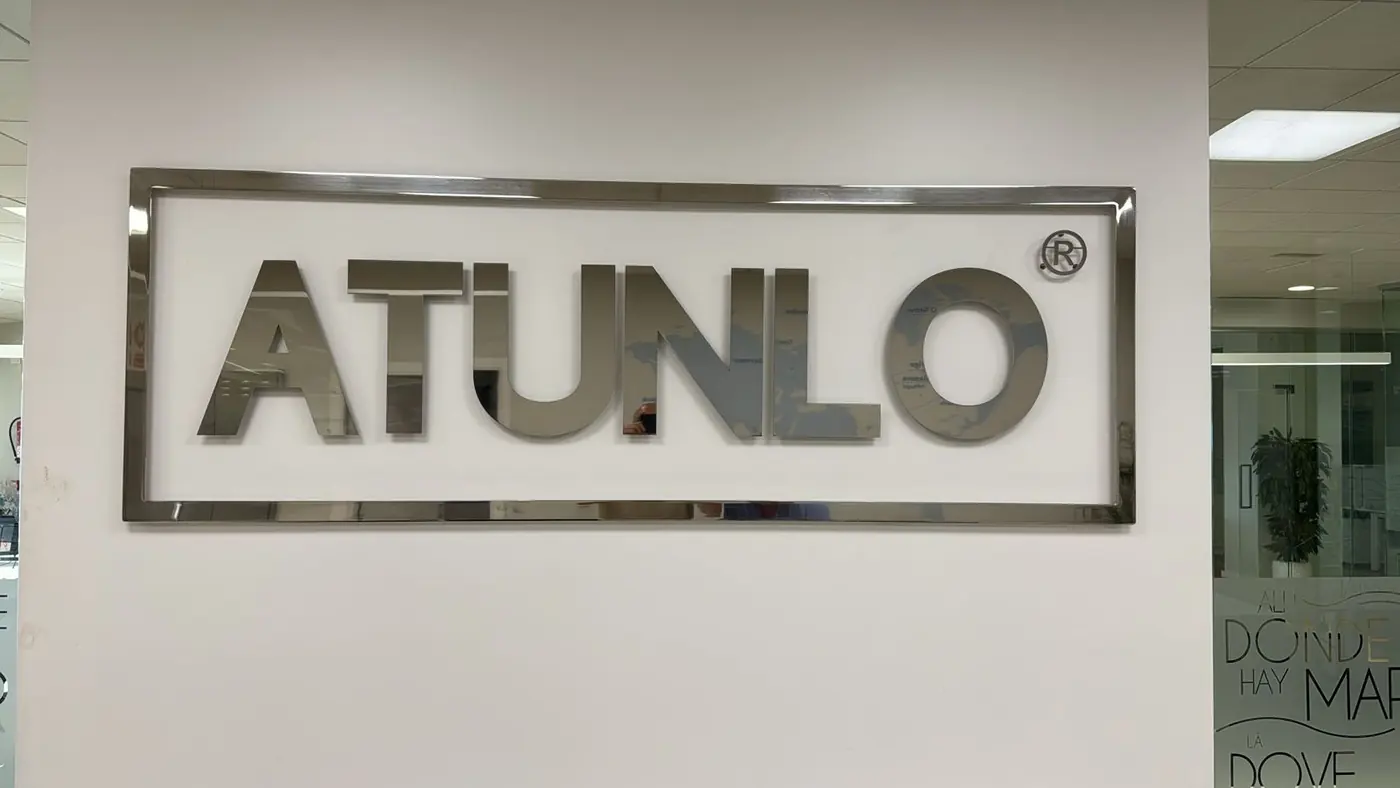 Logotipo de Atunlo en letras de acero pulido dentro de un marco, sobre pared blanca