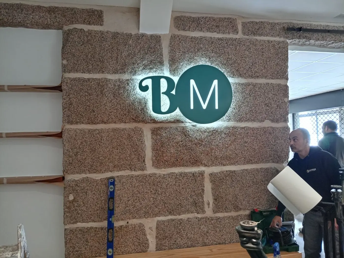 Logotipo BM en letras retroiluminadas sobre pared de piedra, durante la instalación