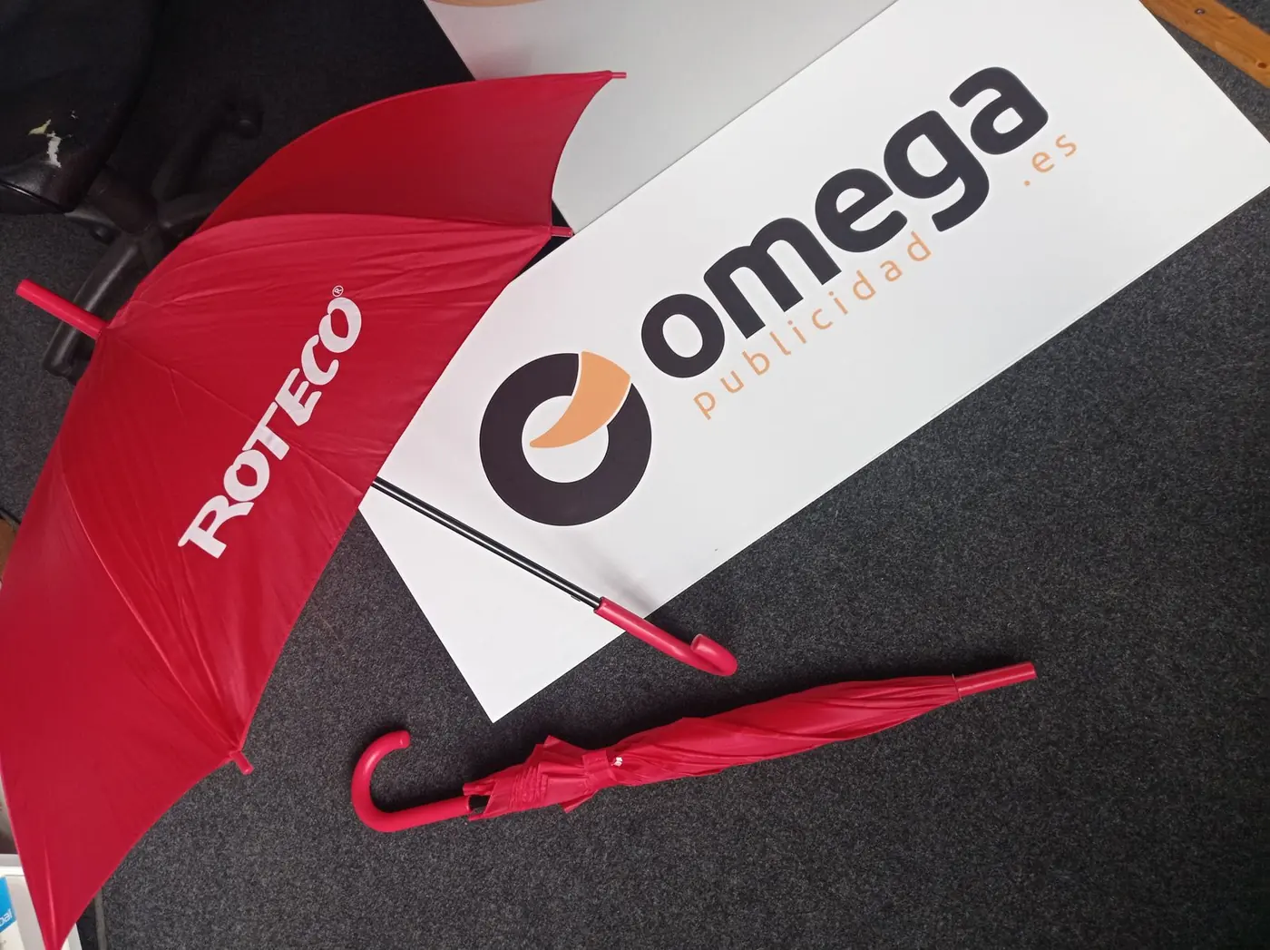 Paraguas rojos con el nombre Roteco, abierto y plegado, junto al logotipo de Omega