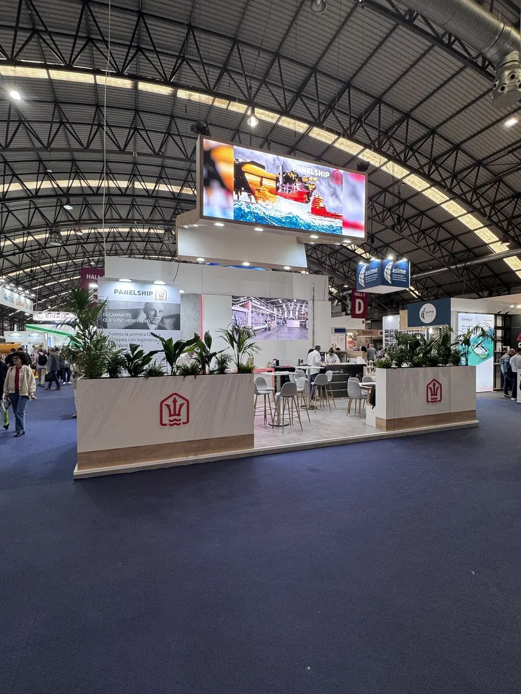 Stand grande en una feria, con estructura blanca y verde bajo la cubierta metálica de la nave